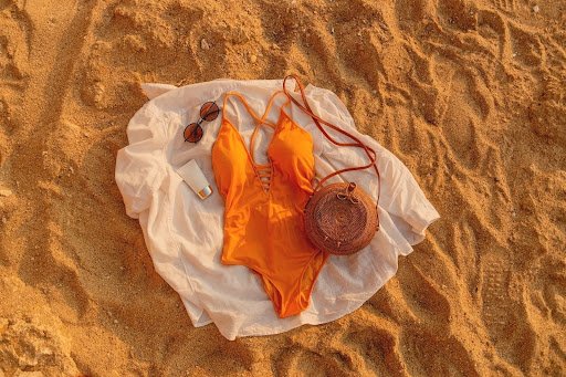 Orange swimsuit with sunglasses, sunscreen, and straw bag laid out on sandy beach.