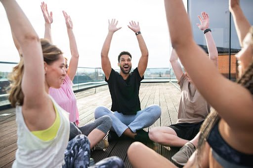 Group of people sitting in a circle on deck outdoors raising arms in a joyful activity.