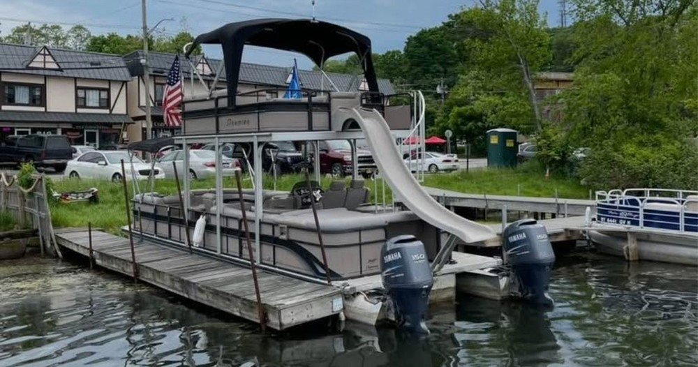 Pontoon boat with water slide docked near lakeshore with twin Yamaha outboard engines.