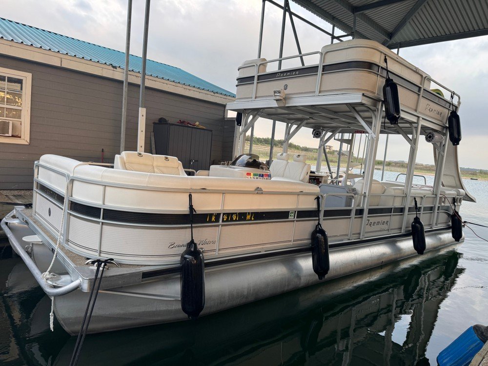 Premier pontoon boat with white seating and upper deck roof parked at marina dock.