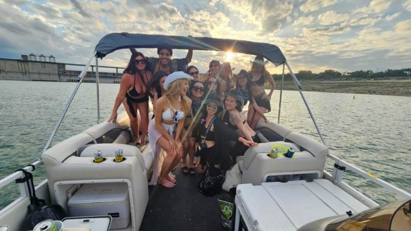roup of women on pontoon boat – A group of women in swimsuits and hats smiling and posing on a pontoon boat with the sunset in the background.