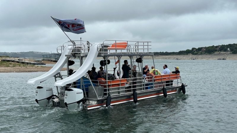 Pontoon boat with waterslide carrying a group of people enjoying a sunny day on the lake.